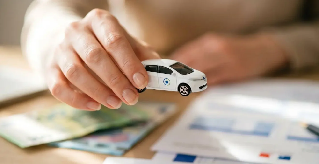 Main tenant une petite voiture électrique miniature avec billets en euros en arrière-plan, symbolisant les aides gouvernementales à l'achat